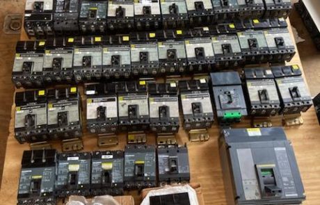 A large assortment of industrial circuit breakers A large assortment of industrial circuit breakers neatly arranged on a wooden table. The breakers vary in size and brand, with most featuring black casings and yellow labels. Some are wrapped in protective packaging. In the background, cardboard boxes and wooden pallets are visible, and a person’s foot can be seen in the top-right corner. The setting appears to be a warehouse or storage area.