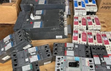 United-Industries-Products-03 A collection of various circuit breakers of different sizes and brands laid out on a wooden table. Some of the breakers are large with lever switches, while others are smaller with push-button or toggle mechanisms. The breakers appear used or refurbished, with visible wear and labels. In the background, there are multiple cardboard boxes, some open, containing additional electrical components and packaging materials.