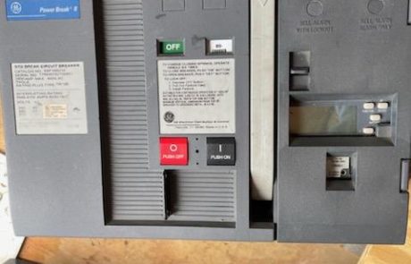 United-Industries-Products-04 A GE Power Break II circuit breaker with a black and gray casing, placed on a wooden surface. The breaker has a red "Push Off" button and a black "Push On" button, along with a green "Off" indicator. Various labels and warnings are visible, including a specifications label on the left side. The breaker appears to be in a used or refurbished condition. In the background, other electrical components are partially visible.
