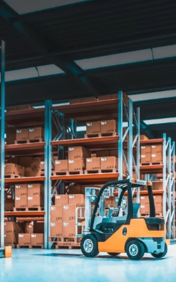 Our Surplus Equipment Liquidation Process A small, yellow forklift sits parked in an industrial warehouse filled with rows of tall shelving units stacked with cardboard boxes, highlighting United Industries' efficient logistics.