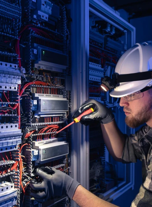 Expert Maintenance of Industrial Electrical Switchgear Panel Electrical technician in a hard hat and safety glasses inspecting a complex industrial switchgear panel with a screwdriver.